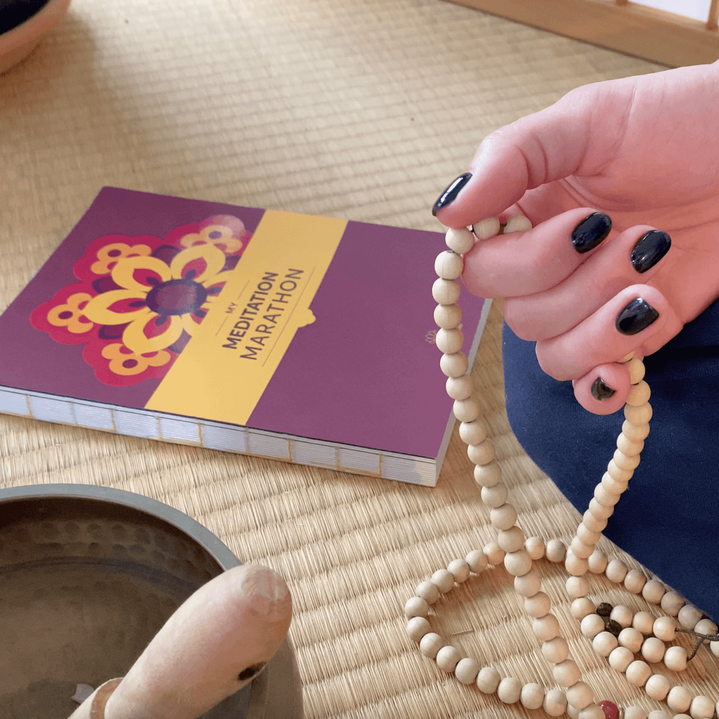 Hand with mandala beads, meditation journal, and singing bowl on a tatami mat.